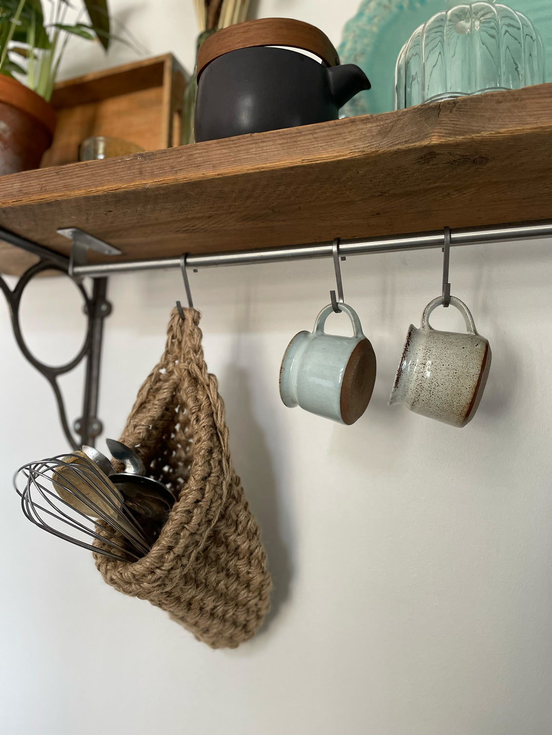 Image showing kitchen shelf with hanging storage bag suspended by a metal hook with handmade storage bag containing kitchen utensils, Jute hanging storage bag holing with whisk and ladle, handmade sustainable crochet decor, rustic natural organic homeware accessories, brown strong jute storage solution, kitchen bathroom bedroom hanging storage bag