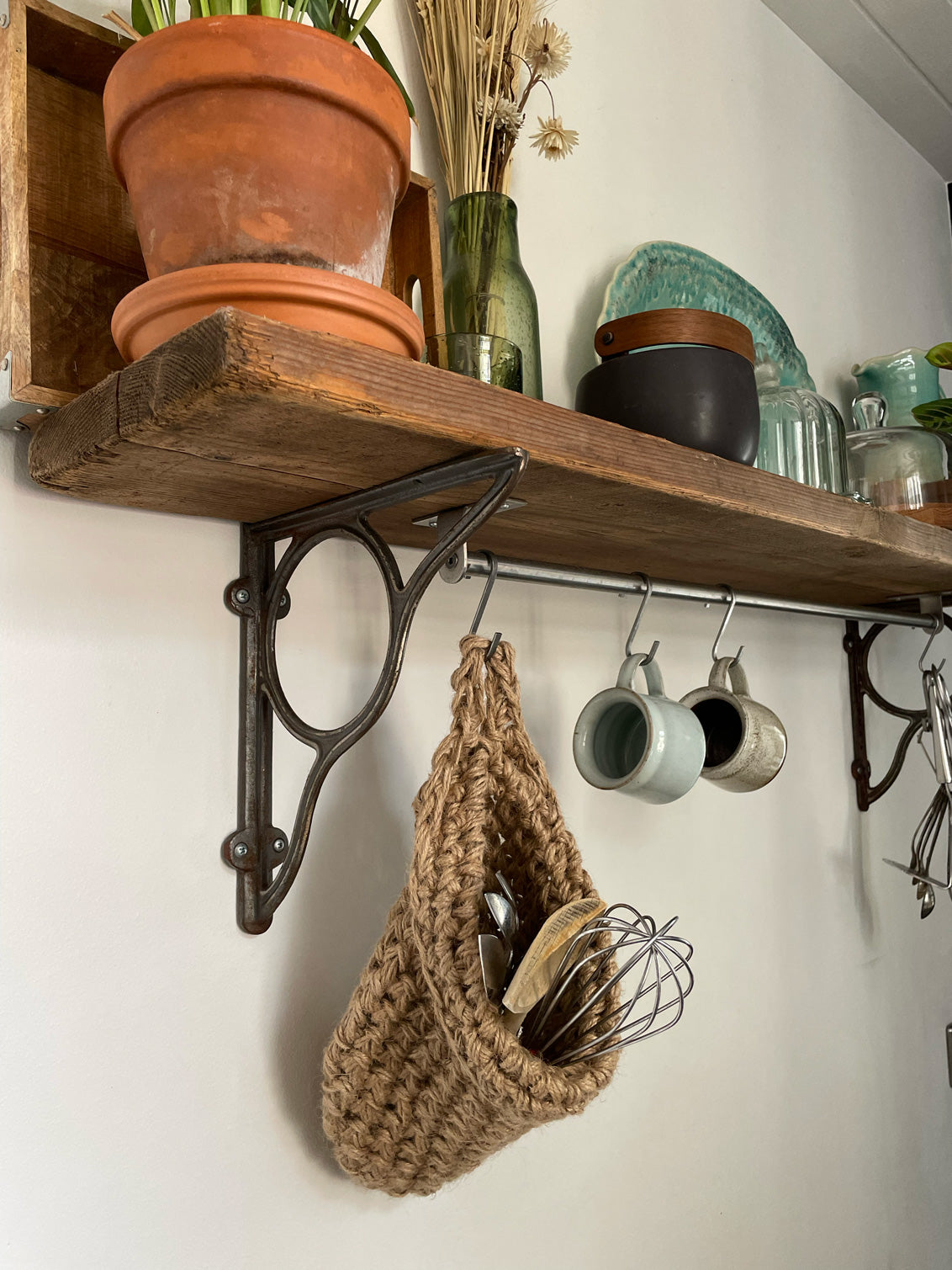 Image showing kitchen shelf with hanging storage bag suspended by a metal hook with handmade storage bag containing kitchen utensils, Jute hanging storage bag holing with whisk and ladle, handmade sustainable crochet decor, rustic natural organic homeware accessories, brown strong jute storage solution, kitchen bathroom bedroom hanging storage bag