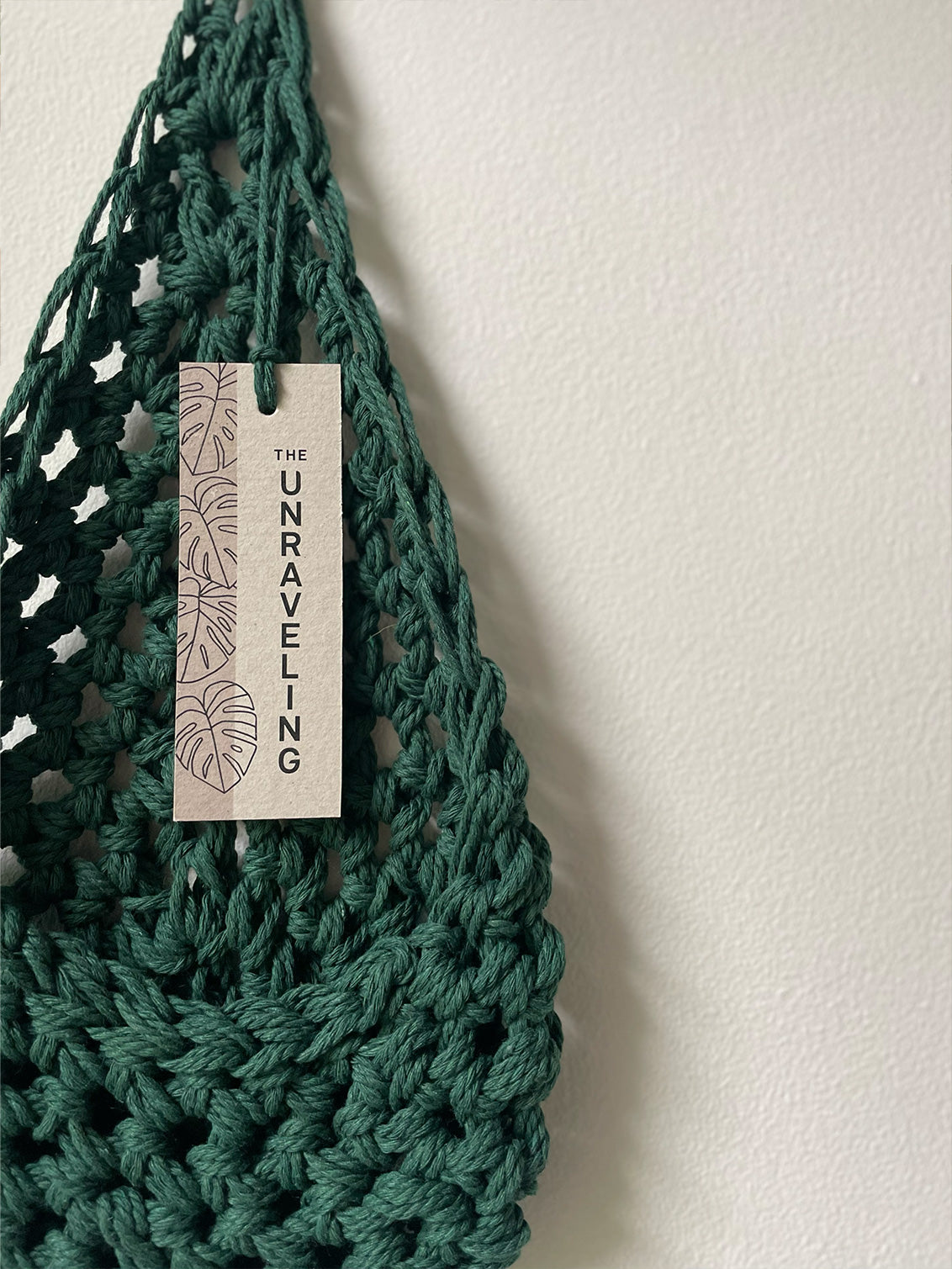 Image showing handmade green cotton planter hanging against a white wall with swing tag attached showing brand name, The Unraveling, Planter is flat to show the tierdrop shape before adding pot and plant.