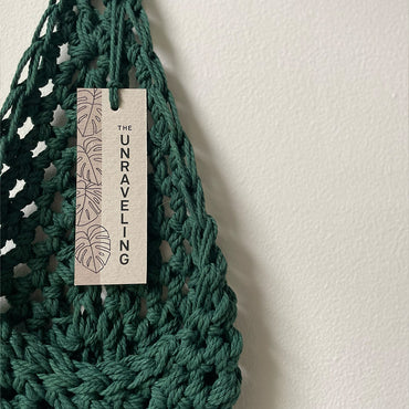 Image showing handmade green cotton planter hanging against a white wall with swing tag attached showing brand name, The Unraveling, Planter is flat to show the tierdrop shape before adding pot and plant.