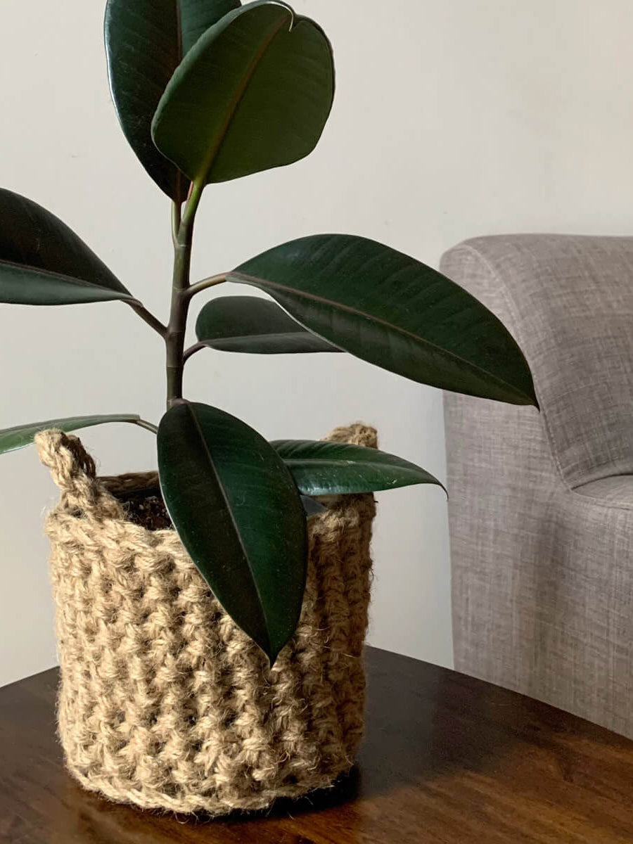 Photo showing crocheted jute plant basket, containing plant, placed on a small wooden table. Placed to the side of a living room sofa. The plant basket is made of natural jute, brown, and has two small handles.
