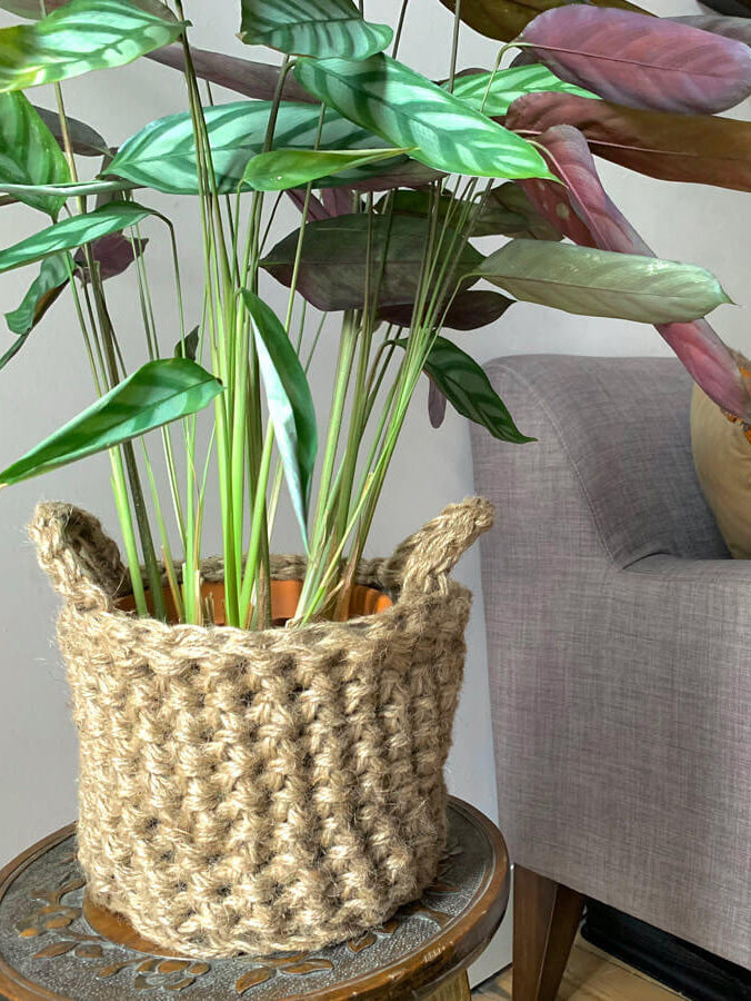 Photo showing crocheted jute plant basket, containing plant, placed on a small wooden table. Placed to the side of a living room sofa. The plant basket is made of natural jute, brown, and has two small handles. 