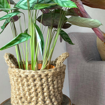 Photo showing crocheted jute plant basket, containing plant, placed on a small wooden table. Placed to the side of a living room sofa. The plant basket is made of natural jute, brown, and has two small handles. 