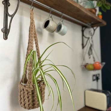 Natural Jute hanging plant holder with three long crocheted straps and pot shaped basket at the bottom of these straps designed for pot to be placed securely, finished with a loop to hang up. Image shows planter hanging in a kitchen, from a hanging rail, with a pot and plant inside.