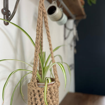 Natural Jute hanging plant holder with three long crocheted straps and pot shaped basket at the bottom of these straps designed for pot to be placed securely, finished with a loop to hang up. Image shows planter hanging in a kitchen, from a hanging rail, with a pot and plant inside.