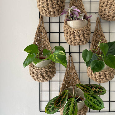 Image showing a close up photo of a grouping of 6 tierdrop shaped natural jute wall hanging planters. Each handmade planter is hung against the wall, holding a ceramic pot and plant. Photo is showing a creative sapce saving method of showing of your houseplants.