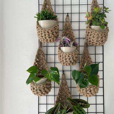 Image showing a grouping of 6 tierdrop shaped natural jute wall hanging planters. Each handmade planter is hung against the wall, holding a ceramic pot and plant. Photo is showing a creative sapce saving method of showing of your houseplants.