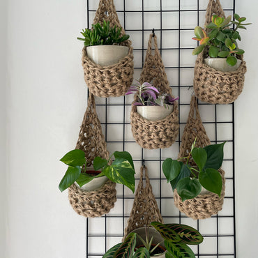 Image showing a grouping of 6 tierdrop shaped natural jute wall hanging planters. Each handmade planter is hung against the wall, holding a ceramic pot and plant. Photo is showing a creative sapce saving method of showing of your houseplants. 