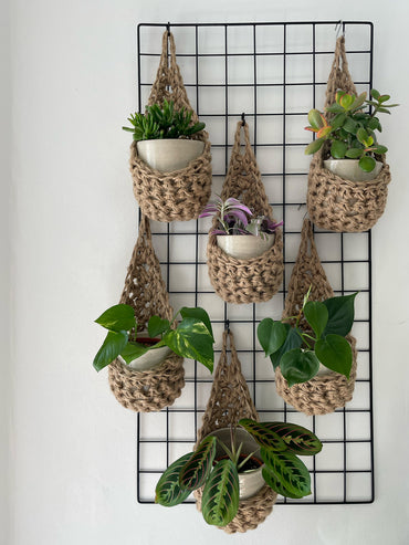 Image showing a grouping of 6 tierdrop shaped natural jute wall hanging planters. Each handmade planter is hung against the wall, holding a ceramic pot and plant. Photo is showing a creative sapce saving method of showing of your houseplants. 
