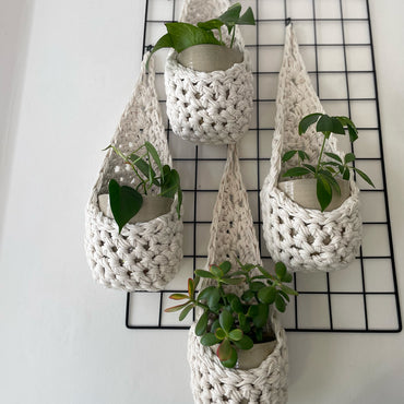 Image of a white wall with four white wall mounted planters hanging together in a group, each one containing a pots and plant. Handmade cotton crochet plant baskets, used to display houseplants creatively in your home. Photo shows the basket shape which keeps your plants safe and secure