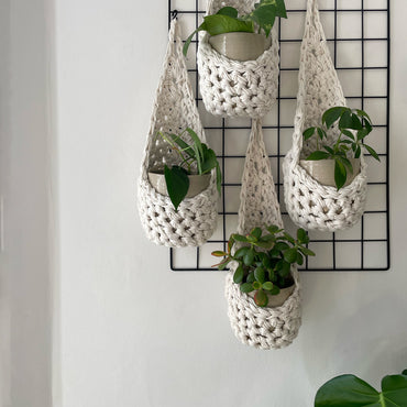 Image of a white wall with four white wall mounted planters hanging together in a group, each one containing a pots and plant. Handmade cotton crochet plant baskets, used to display houseplants creatively in your home. Photo shows the basket shape which keeps your plants safe and secure