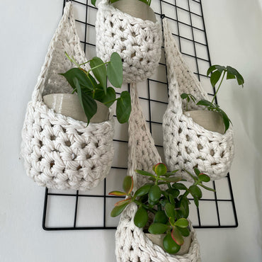 Image showing low angled shot of a white wall with four white wall mounted planters hanging together in a group, each one containing a pots and plant. Handmade cotton crochet plant baskets, used to display houseplants creatively in your home. Photo shows the basket shape which keeps your plants safe and secure.