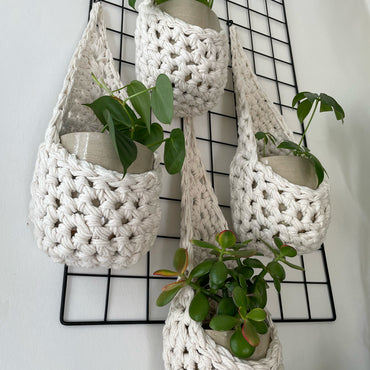 Image showing low angled shot of a white wall with four white wall mounted planters hanging together in a group, each one containing a pots and plant. Handmade cotton crochet plant baskets, used to display houseplants creatively in your home. Photo shows the basket shape which keeps your plants safe and secure. 