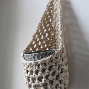 Image of one ivory cotton crochet hanging wall planter against a white wall. The photo shows the planter with a pot, without a plant, in order to demonstate tierdrop shape of design.