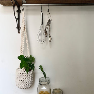 Indoor cream cotton hanging plant holder with three long crocheted straps, finished with a loop to hang up. Image shows planter hanging in a kitchen, from a hanging rail, with a pot and plant inside. Made with recycled cotton cord.