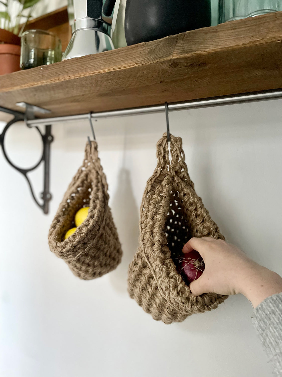 mage showing kitchen shelf with hanging storage bag suspended by a metal hook, handmade storage bag containing red onions in a kitchen, breathable storage bag for kitchen produce displayed on a hanging rail, space saving hanging storage in a kitchen wall, brown jute handmade storage bag