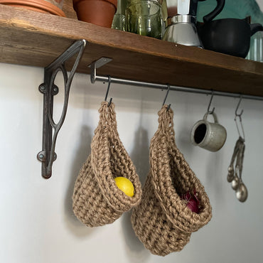 small and large brown jute hanging storage baskets in a kitchen holding lemons and onions, shelf with hooks to hang natural jute storage bags, handmade sustainable crochet decor, rustic natural organic homeware accessories, brown strong jute storage solution, kitchen bathroom bedroom hanging storage bag