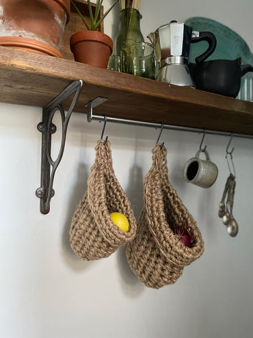small and large brown jute hanging storage baskets in a kitchen holding lemons and onions, shelf with hooks to hang natural jute storage bags, handmade sustainable crochet decor, rustic natural organic homeware accessories, brown strong jute storage solution, kitchen bathroom bedroom hanging storage bag