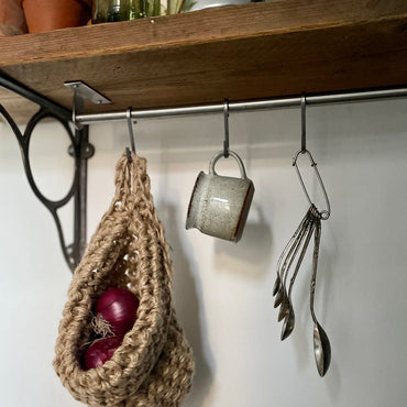 image showing kitchen shelf with hanging storage bag suspended by a metal hook, handmade storage bag containing red onions in a kitchen, breathable storage bag for kitchen produce displayed on a hanging rail, space saving hanging storage in a kirchen wall, brown jute handmade storage bag