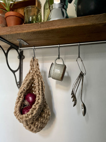 image showing kitchen shelf with hanging storage bag suspended by a metal hook, handmade storage bag containing red onions in a kitchen, breathable storage bag for kitchen produce displayed on a hanging rail, space saving hanging storage in a kirchen wall, brown jute handmade storage bag