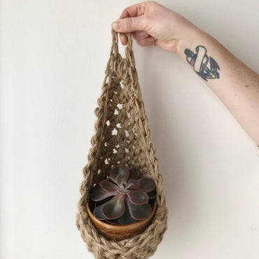 Photo showing a single handmade natural jute wall planter, being held front on by makers hand. Showing it's teardrop shape with ceramic pot and plant inside it.