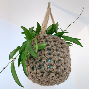 Natural Jute hanging plant holder with three long crocheted straps and pot shaped basket at the bottom of these straps designed for pot to be placed securely, finished with a loop to hang up. Photo showing the view from underneath the basket, showing the basket shape and texture of the firbres. The plant hanger is suspended from the ceiling.