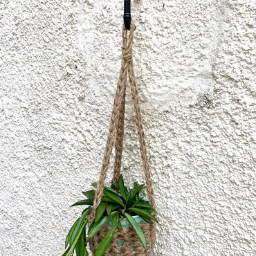 Natural Jute hanging plant holder with three long crocheted straps and pot shaped basket at the bottom of these straps designed for pot to be placed securely, finished with a loop to hang up. Image shows planter outside, suspended from a small hook.