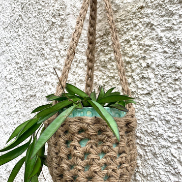 Natural Jute hanging plant holder with three long crocheted straps and pot shaped basket at the bottom of these straps designed for pot to be placed securely, finished with a loop to hang up. Image shows planter outside, suspended from a small hook.