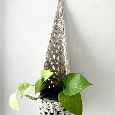 Image of single white cotton crochet hanging wall planter against a wall of a bedroom. The photo shows the planter with a pot and plant inside it, in order to demonstate tierdrop shape of design. Hanging by a small nail in the wall.