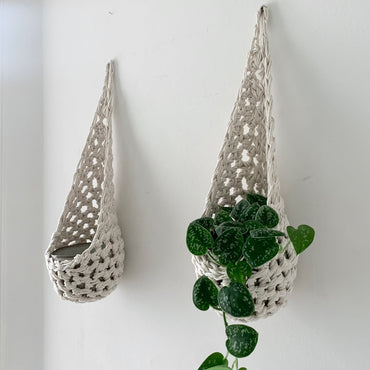 Image of two white cotton crochet hanging wall planter against a wall of a bedroom. The photo shows the planter with a pot and plant inside it, in order to demonstate tierdrop shape of design. Hanging by a small nail in the wall.