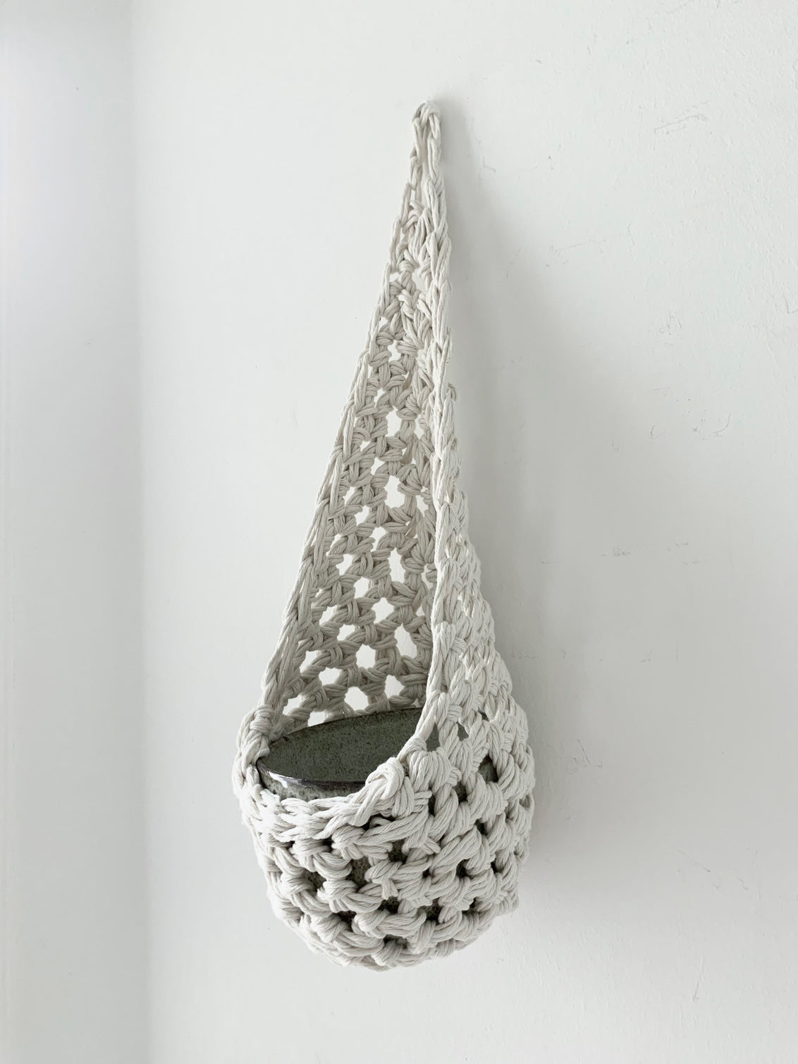 Image of single white cotton crochet hanging wall planter against a wall of a bedroom. The photo shows the planter with a cermaic pot, in order to demonstate tierdrop shape of design. Hanging by a small nail in the wall.