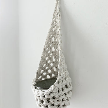 Image of single white cotton crochet hanging wall planter against a wall of a bedroom. The photo shows the planter with a cermaic pot, in order to demonstate tierdrop shape of design. Hanging by a small nail in the wall.