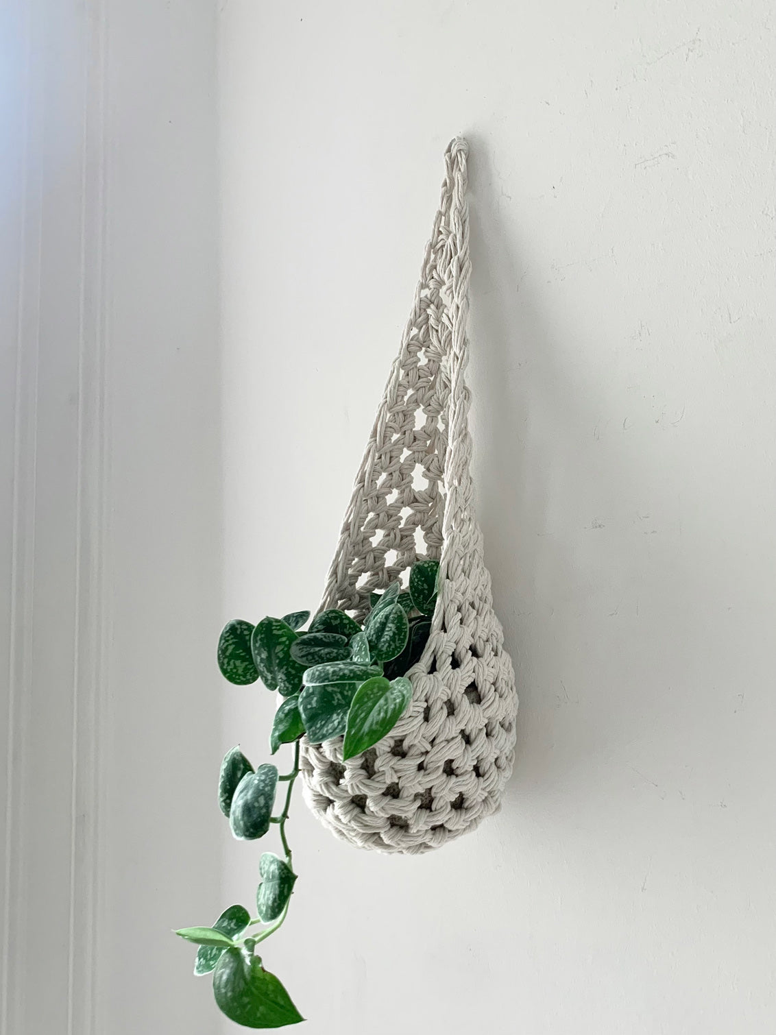 Image of single white cotton crochet hanging wall planter against a wall of a bedroom. The photo shows the planter with a pot and plant inside it, in order to demonstate tierdrop shape of design. Hanging by a small nail in the wall.
