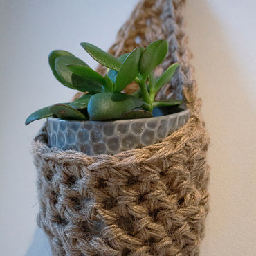 Photo showing a single handmade crochet jute hanging wall planter, against a white wall. The planter is a tierdrop shape and is holding a pot and plant