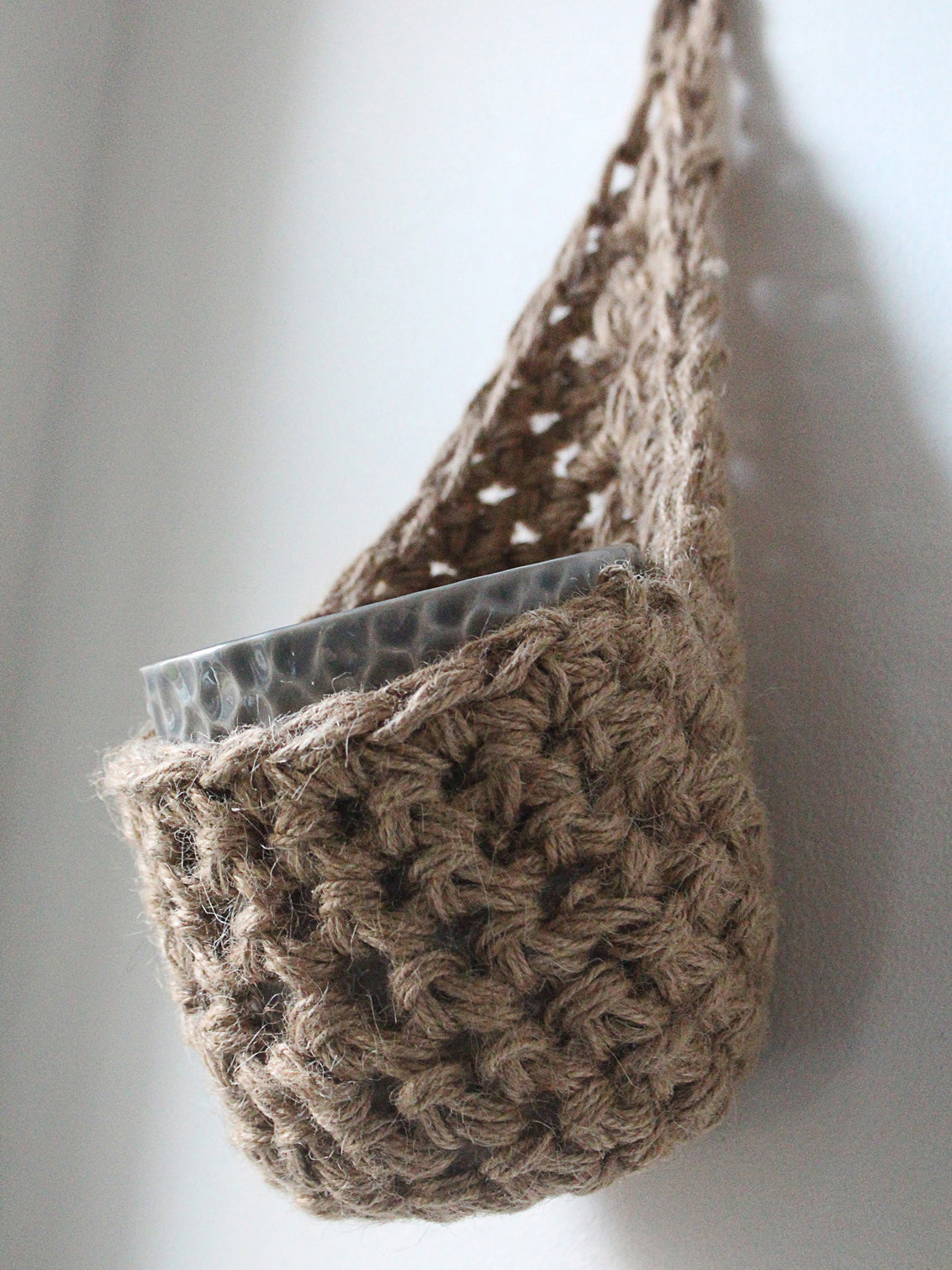 Photo showing a single handmade crochet jute hanging wall planter, against a white wall. The planter is a tierdrop shape and is holding a pot only.