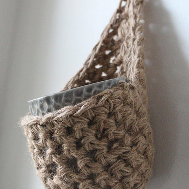 Photo showing a single handmade crochet jute hanging wall planter, against a white wall. The planter is a tierdrop shape and is holding a pot only.