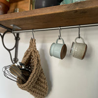Image showing kitchen shelf with hanging storage bag suspended by a metal hook with handmade storage bag containing kitchen utensils, Jute hanging storage bag holing with whisk and ladle, handmade sustainable crochet decor, rustic natural organic homeware accessories, brown strong jute storage solution, kitchen bathroom bedroom hanging storage bag