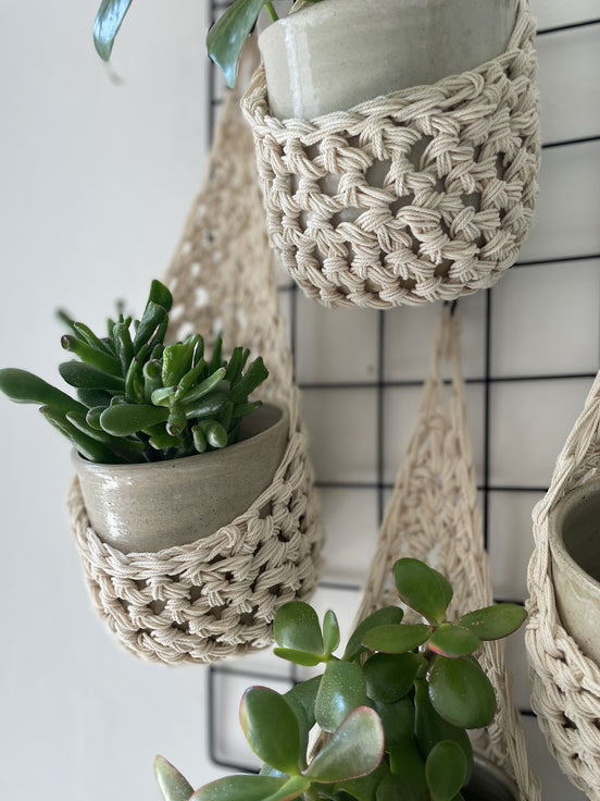 Image showing zoomed in details of four cream wall mounted planters hanging together in a group, each one containing a pots and plant. Handmade cotton crochet plant baskets, used to display houseplants creatively in your home. 