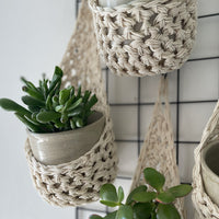 Image showing zoomed in details of four cream wall mounted planters hanging together in a group, each one containing a pots and plant. Handmade cotton crochet plant baskets, used to display houseplants creatively in your home. 
