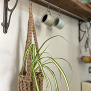 Natural Jute hanging plant holder with three long crocheted straps and pot shaped basket at the bottom of these straps designed for pot to be placed securely, finished with a loop to hang up. Image shows planter hanging in a kitchen, from a hanging rail, with a pot and plant inside.