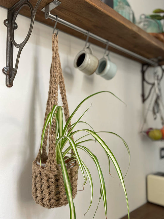 Natural Jute hanging plant holder with three long crocheted straps and pot shaped basket at the bottom of these straps designed for pot to be placed securely, finished with a loop to hang up. Image shows planter hanging in a kitchen, from a hanging rail, with a pot and plant inside.