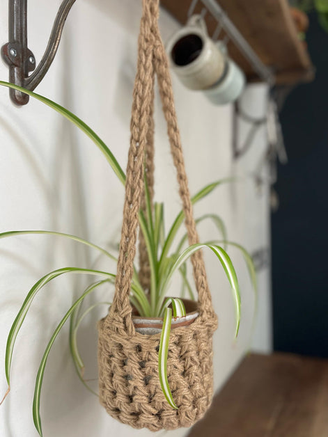 Natural Jute hanging plant holder with three long crocheted straps and pot shaped basket at the bottom of these straps designed for pot to be placed securely, finished with a loop to hang up. Image shows planter hanging in a kitchen, from a hanging rail, with a pot and plant inside.