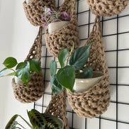 Image showing a close up photo of a grouping of 6 tierdrop shaped natural jute wall hanging planters. Each handmade planter is hung against the wall, holding a ceramic pot and plant. Photo is showing a creative sapce saving method of showing of your houseplants.  