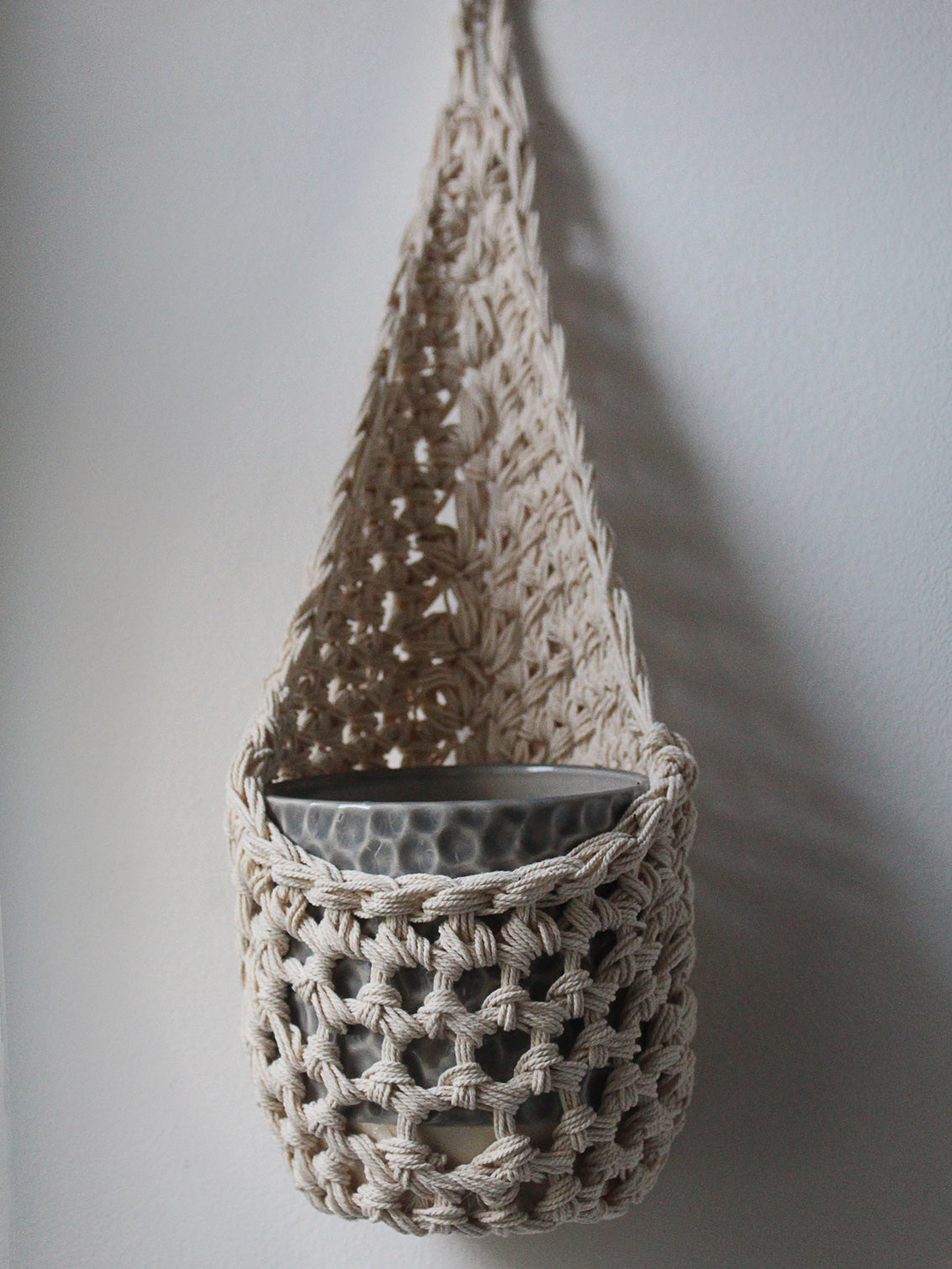 Image of one ivory cotton crochet hanging wall planter against a white wall. The photo shows the planter with a pot, without a plant, in order to demonstate tierdrop shape of design.
