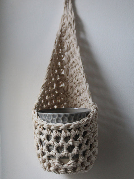 Image of one ivory cotton crochet hanging wall planter against a white wall. The photo shows the planter with a pot, without a plant, in order to demonstate tierdrop shape of design.