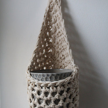 Image of one ivory cotton crochet hanging wall planter against a white wall. The photo shows the planter with a pot, without a plant, in order to demonstate tierdrop shape of design.