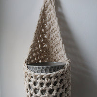 Image of one ivory cotton crochet hanging wall planter against a white wall. The photo shows the planter with a pot, without a plant, in order to demonstate tierdrop shape of design.