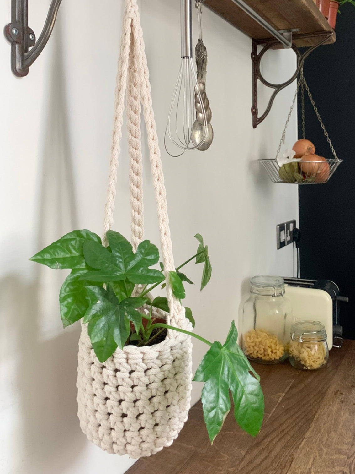 Indoor cream cotton hanging plant holder with three long crocheted straps, finished with a loop to hang up. Image shows planter hanging in a kitchen, from a hanging rail, with a pot and plant inside. Made with recycled cotton cord.