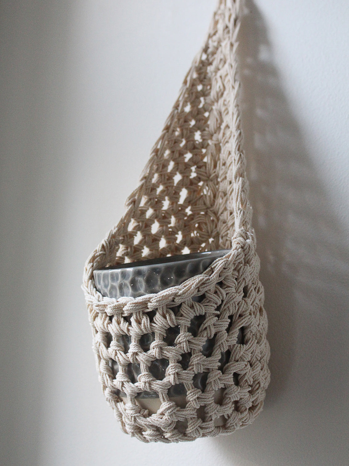 Image of one ivory cotton crochet hanging wall planter against a white wall. The photo shows the planter with a pot, without a plant, in order to demonstate tierdrop shape of design.