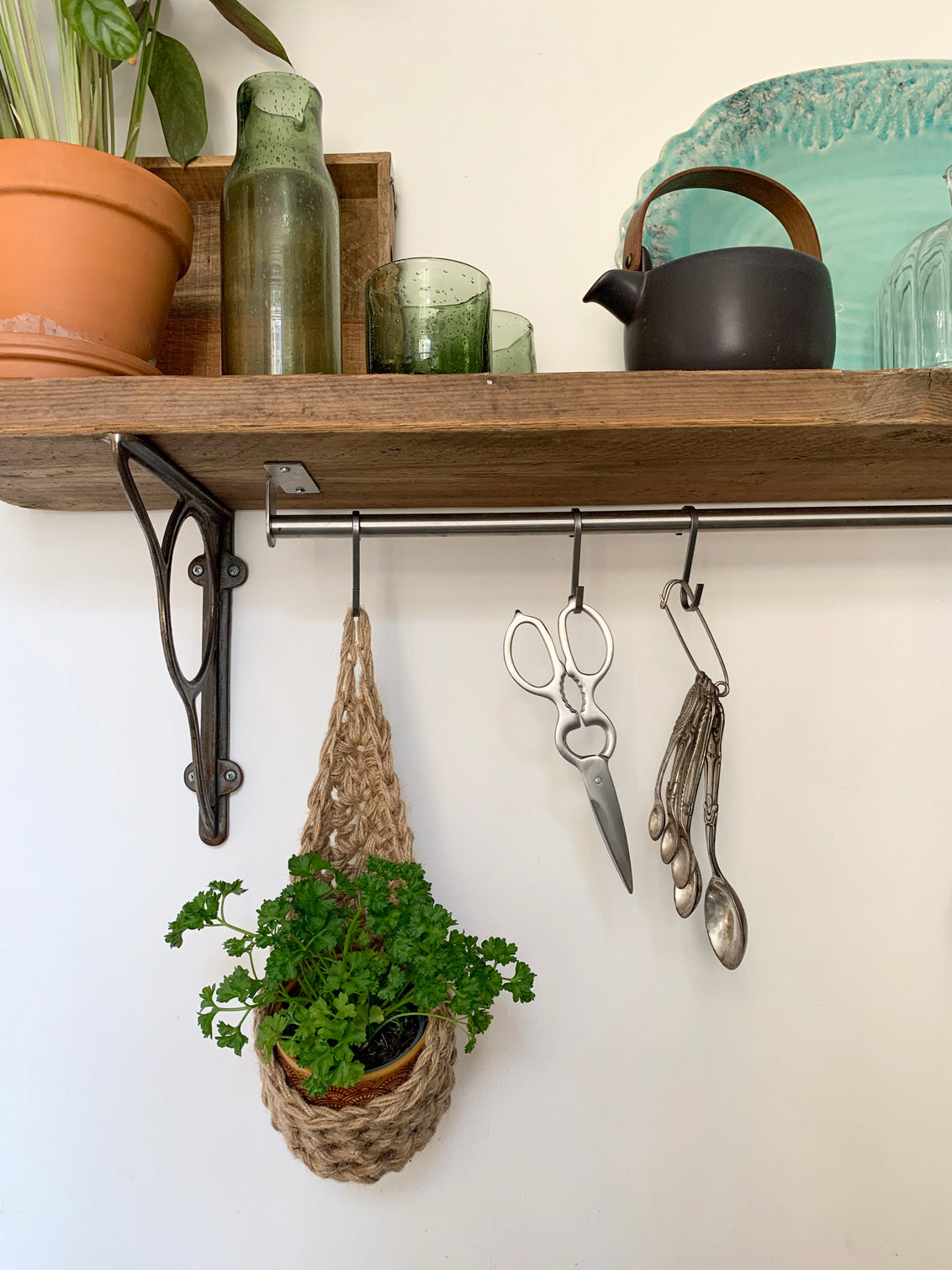 Photo showing small jute hanging planter in a kitchen, holding a ceramic pot and parsely plant. Planter is hanging from a metal hook attached from a metal rail. 